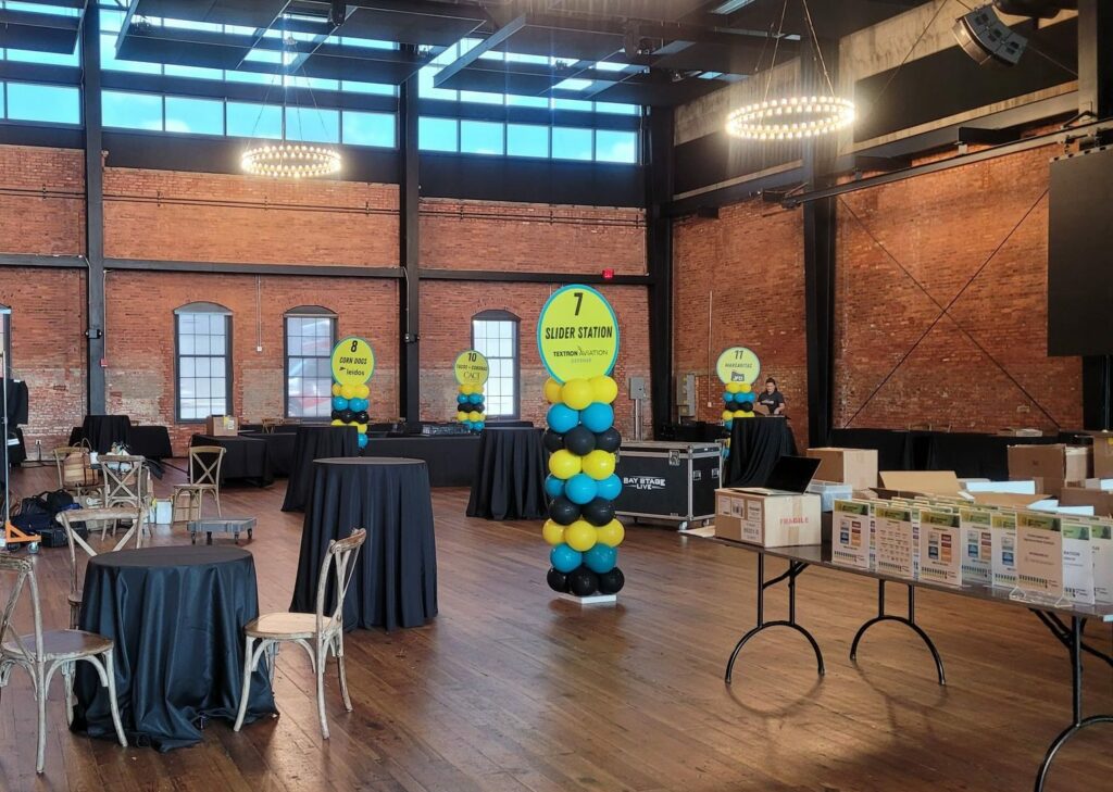 open room with brick walls, tables, chairs, and balloon columns in blue yellow and black directing guests where each station is located