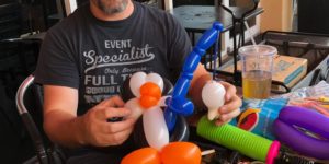 Mr Fudge making a balloon animal penguin with a blue fishing pole catching a white balloon fish, he is sitting at a table