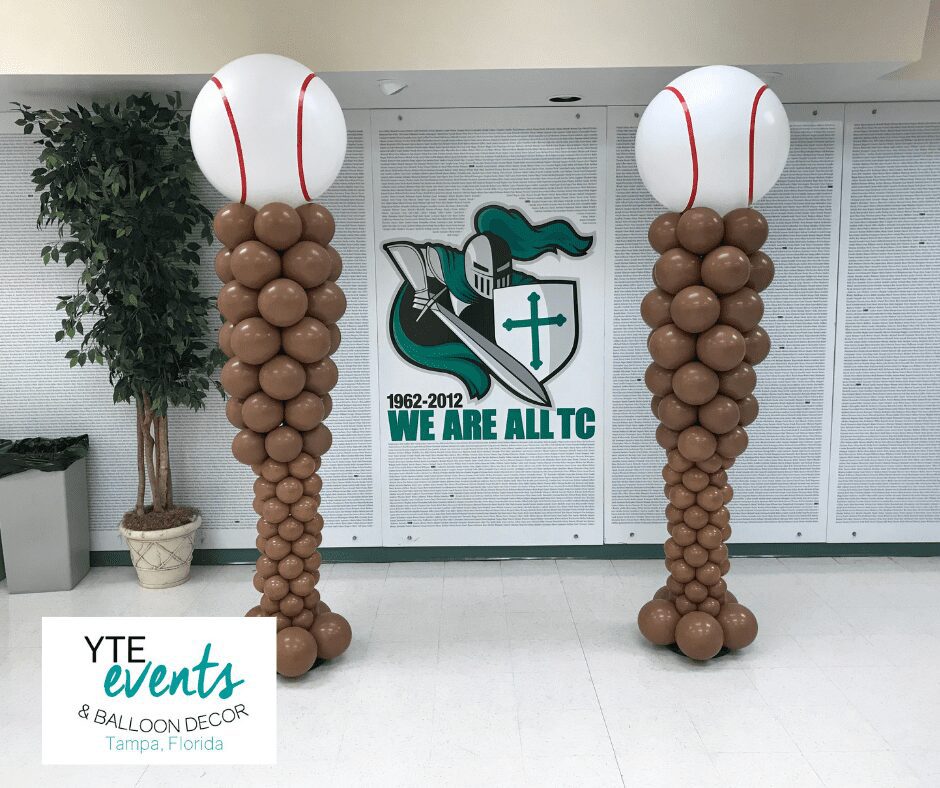 A pair of brown balloon columns with a white baseball balloon topper in a college hallway.