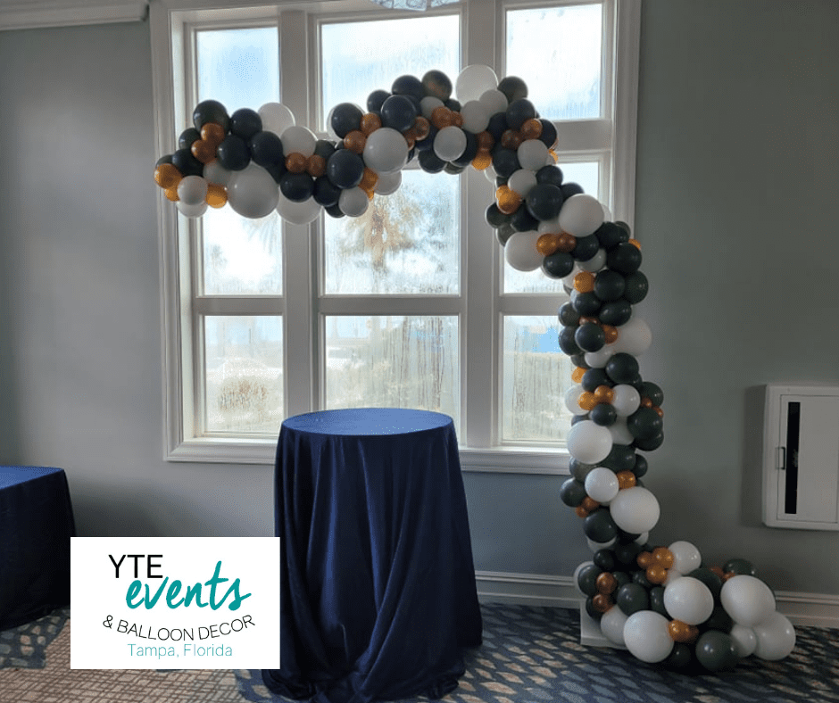 Green gold and white organic balloon decor above a table with a blue tablecloth and in front of a window.