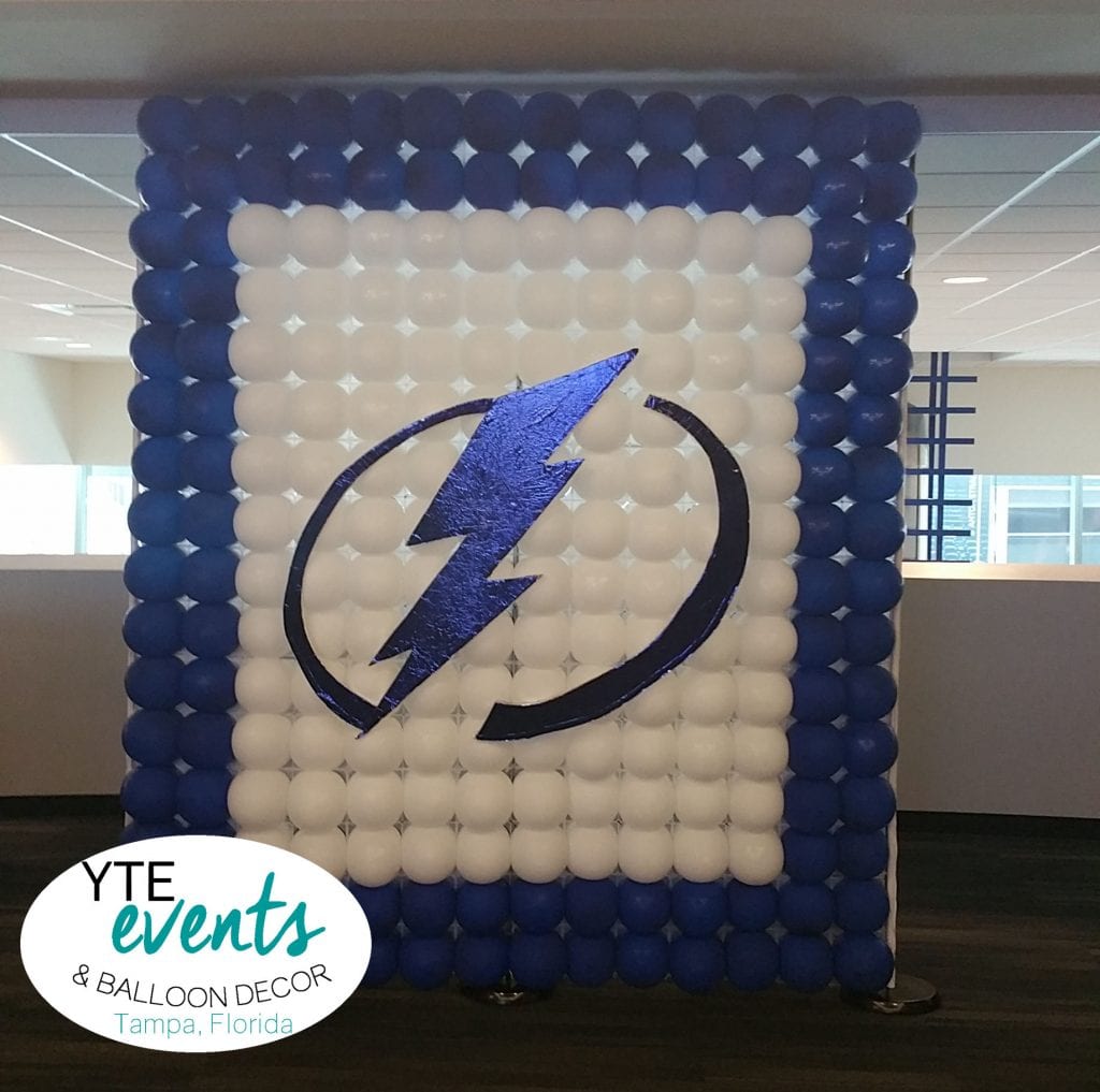 A balloon backdrop created for a Tampa Bay Lightning-themed event. The backdrop consists of a large rectangular structure made entirely of balloons. The outer edges are framed with dark blue balloons, while the inner section is filled with white balloons. In the center, a large blue lightning bolt symbol, the logo of the Tampa Bay Lightning, is prominently displayed. The background shows a modern, well-lit indoor space, suggesting it is set up in a venue such as Amalie Arena in Tampa, Florida. The YTE Events & Balloon Decor logo is visible in the lower left corner, indicating the decorators responsible for this impressive display.