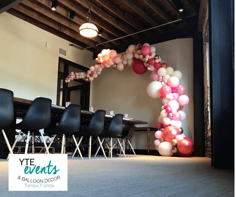 Organic balloon decor made of red pink and white balloons next to a sunny window and a table with chairs.