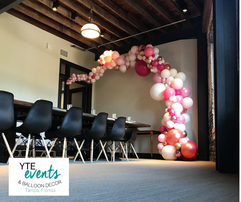 Organic balloon decor made of red pink and white balloons next to a sunny window and a table with chairs.