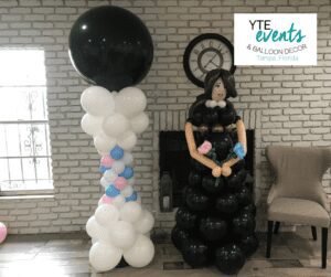 White, pink and blue balloon column with a black balloon topper adjacent to a balloon sculpture of a woman in a black dress with blue and pink flowers.