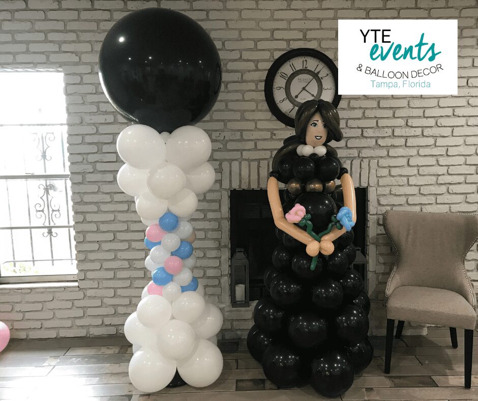 White pink and blue balloon column with a black balloon topper adjacent to a balloon sculpture of a woman in a black dress with blue and pink flowers.