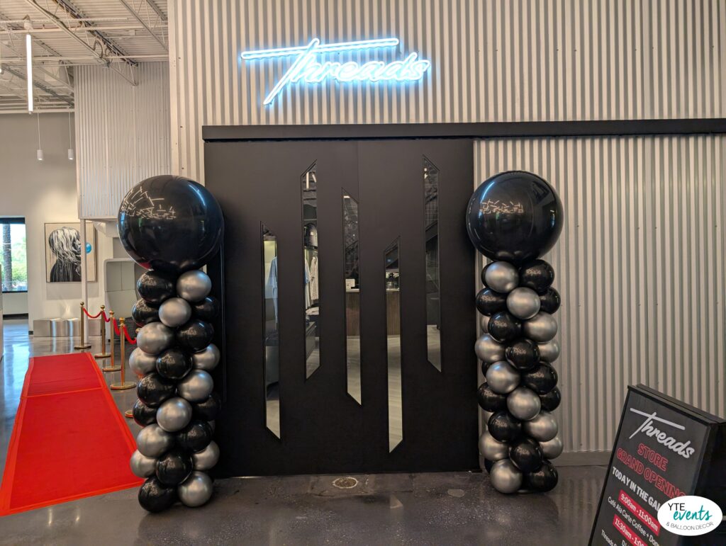 Black and silver balloon columns flanking black doors with Threads neon sign at corporate grand opening event