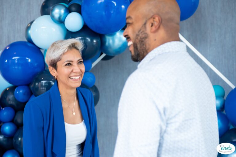 Smiling woman and man in front of a blue balloon arch photobooth at a corporate event