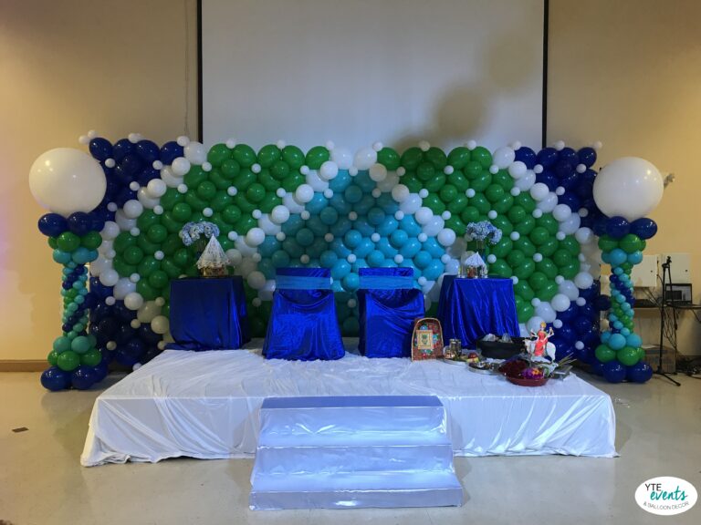 Blue, green, and white balloon backdrop with matching balloon columns, blue draped chairs and tables on a white stage