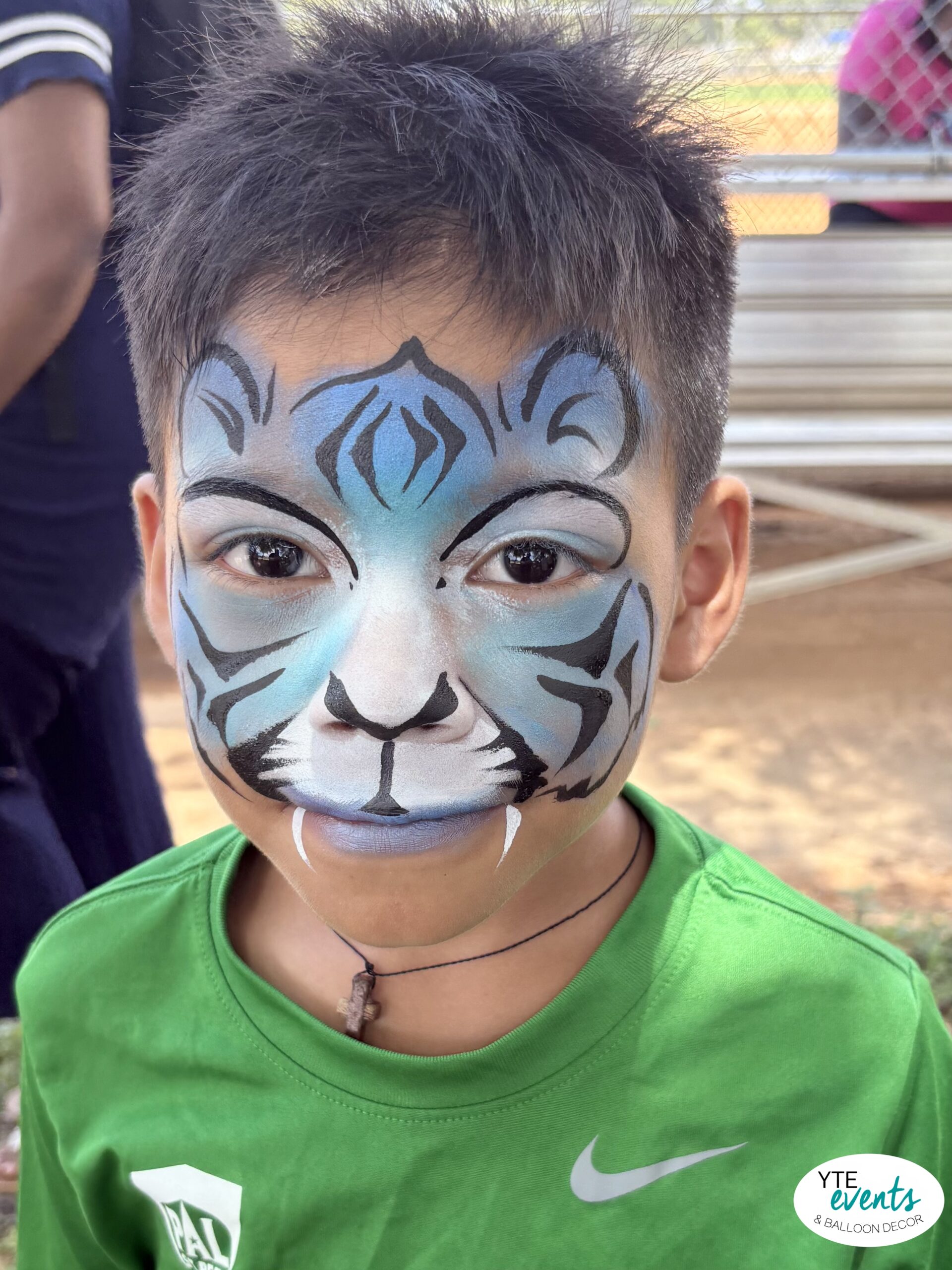 Child with blue tiger face painting wearing green shirt at outdoor kids birthday party