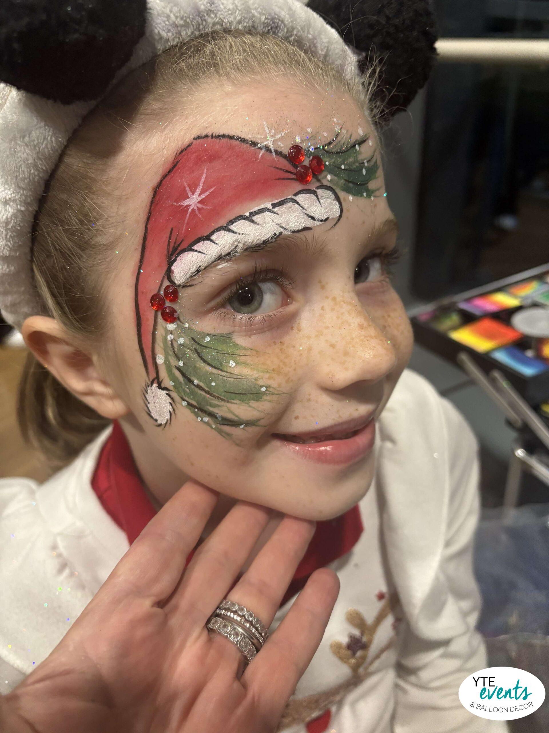Child with Christmas-themed face painting featuring Santa hat and festive greenery