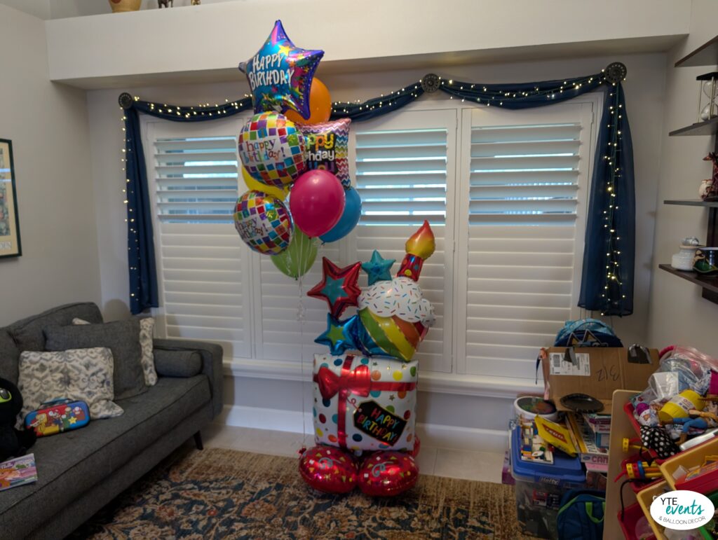 Colorful happy birthday balloon bouquet featuring star-shaped, round, and cake-shaped balloons in a cozy living room.