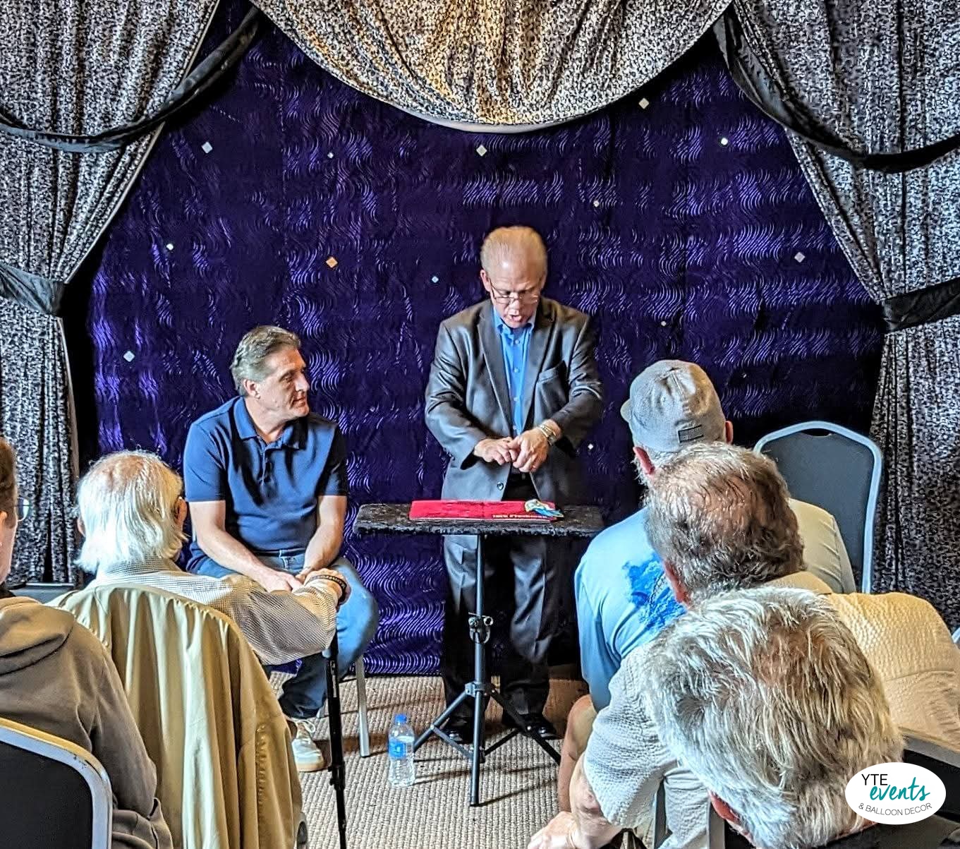 Magician performing at a corporate event with blue fabric backdrop and seated audience
