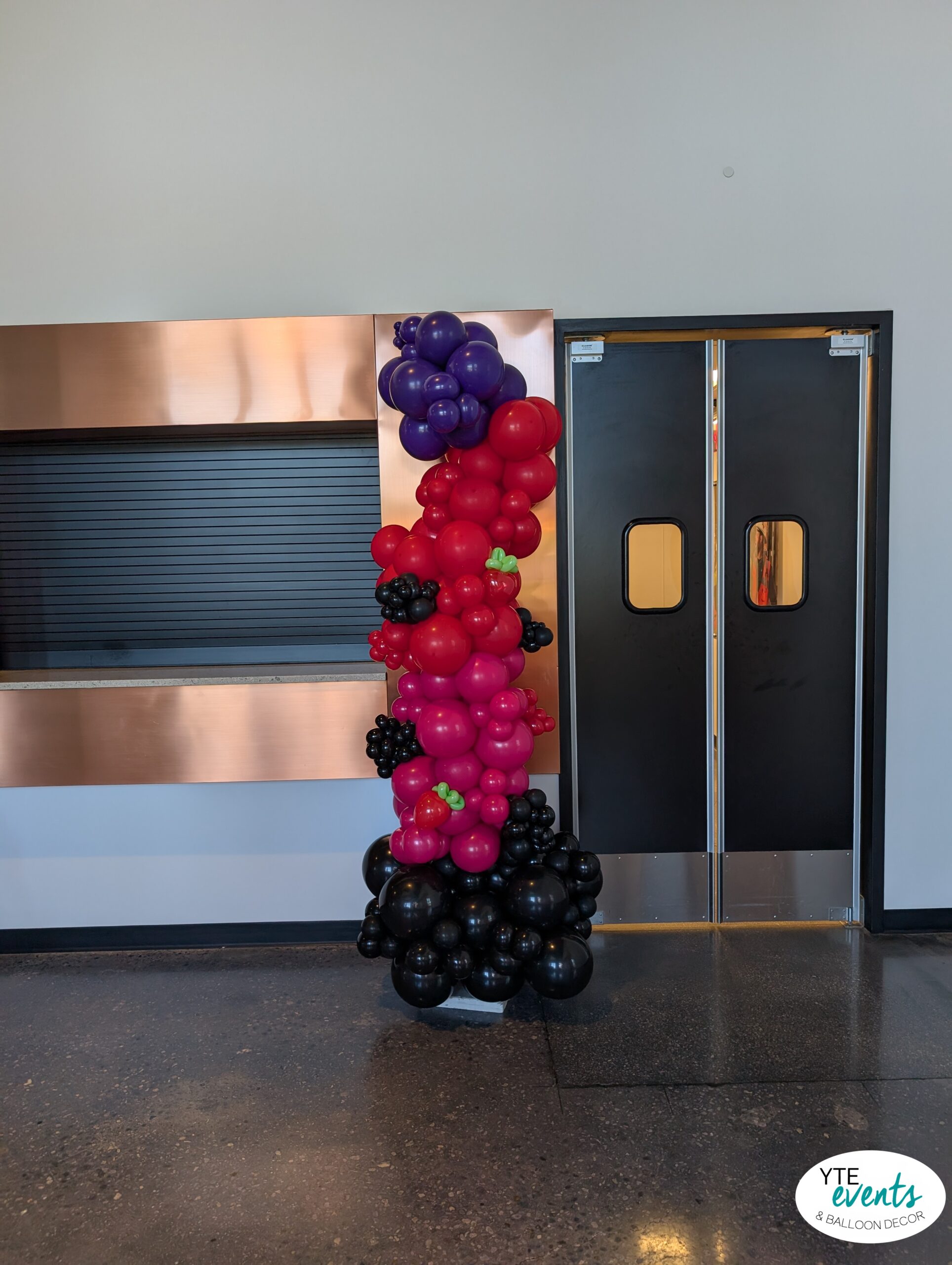 Custom balloon column with black, purple, red, and pink balloons, accented with green leaf-shaped balloons, set indoors near black double doors.