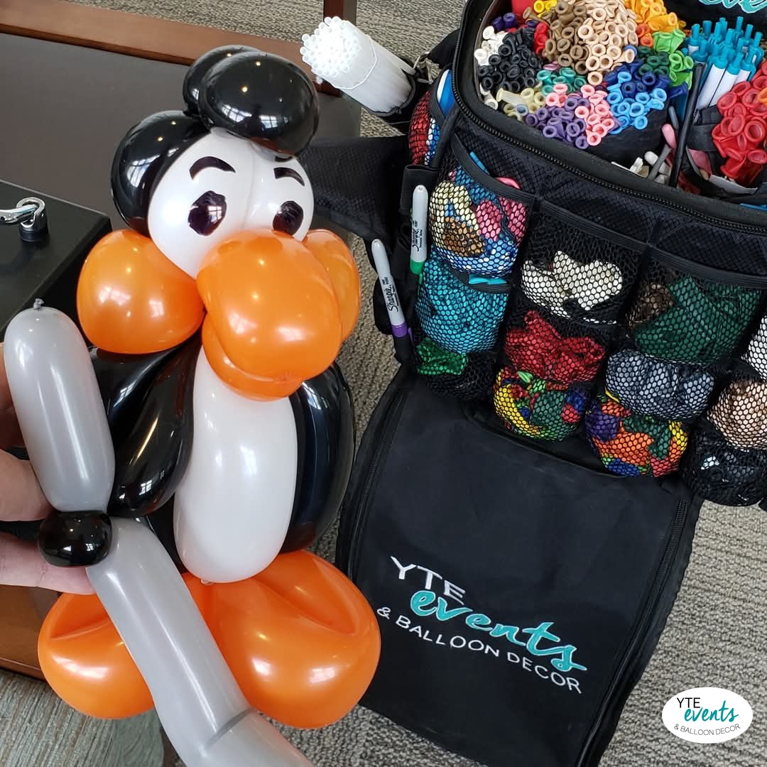 Custom penguin balloon sculpture holding a baseball bat near a balloon decor kit with colorful balloons and markers