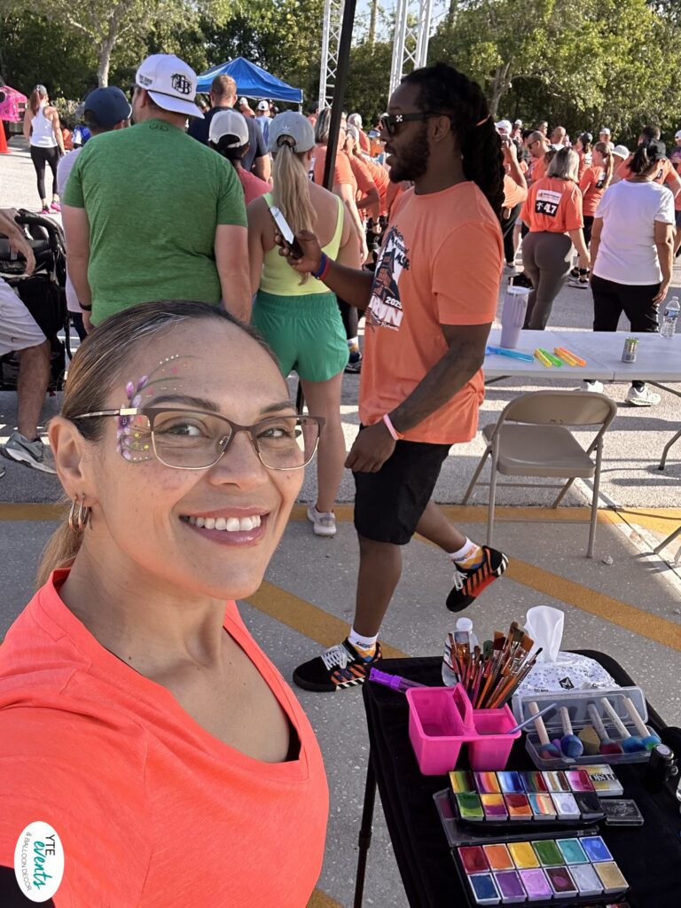Smiling woman with floral face paint design at outdoor community event with crowd and face painting supplies