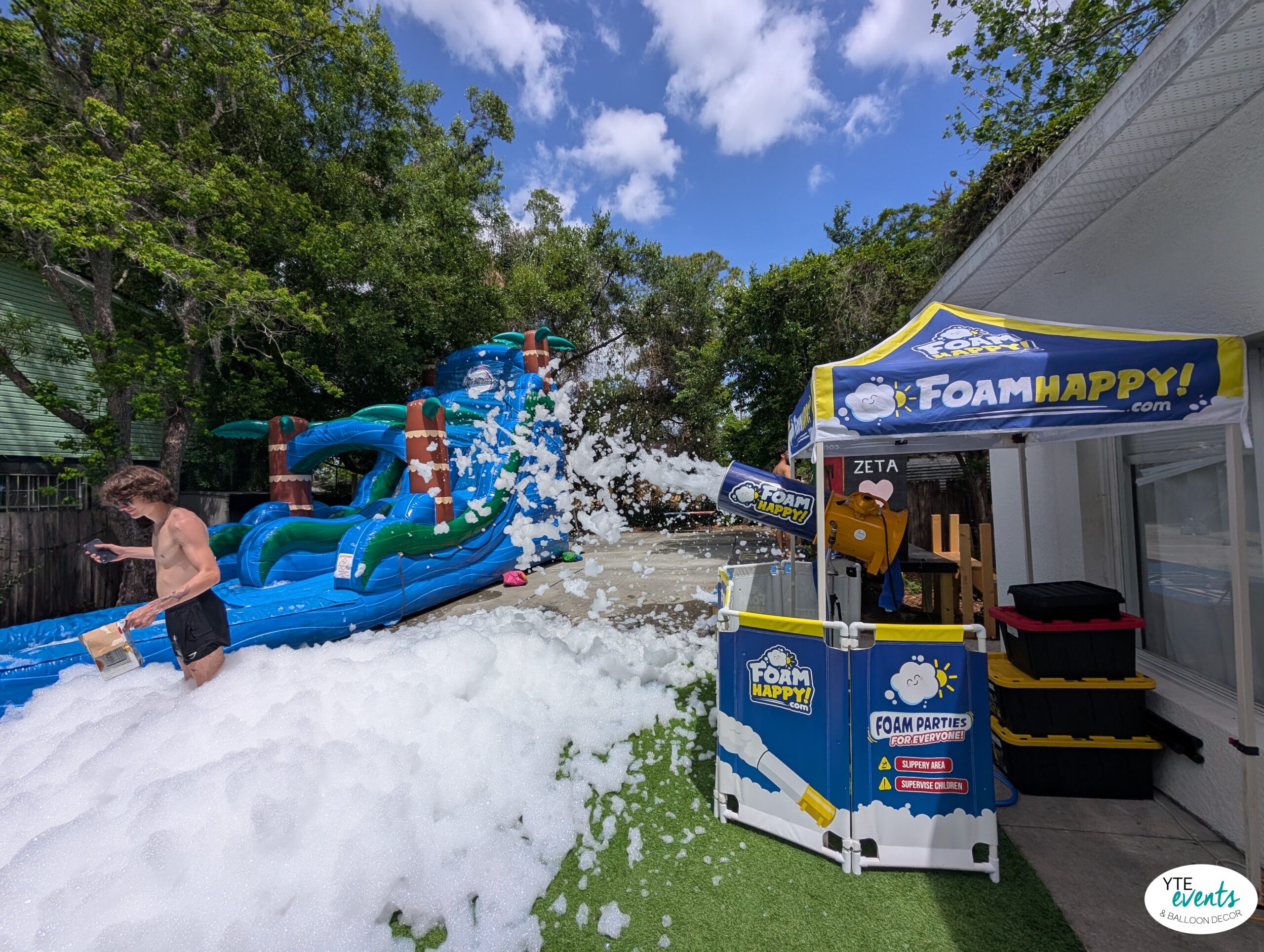 Backyard foam party with blue inflatable water slide and Foam Happy foam machine on a sunny day