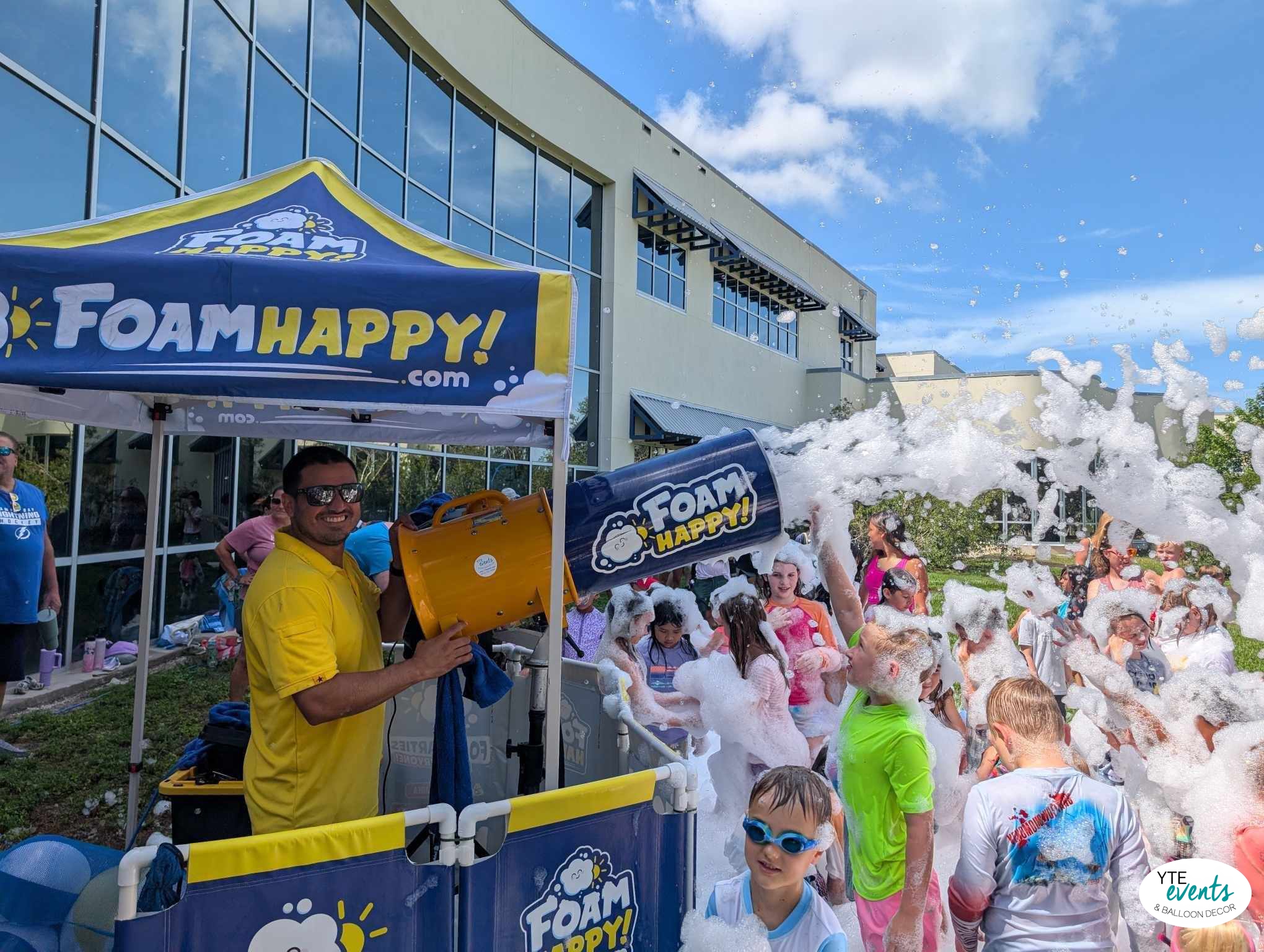Man operating a foam machine creating foam bubbles for children playing outside at a company event