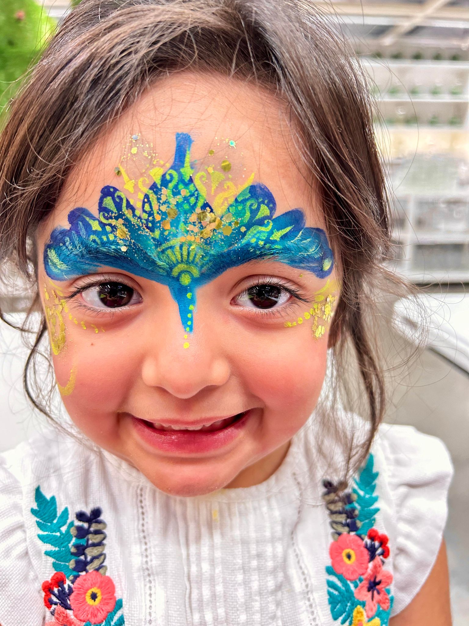 A young girl with intricate face painting featuring a blue and yellow peacock design, extending across her forehead and around her eyes. She is smiling brightly, showcasing the detailed artwork. The setting suggests a family-friendly event or festival in Brandon, Florida.