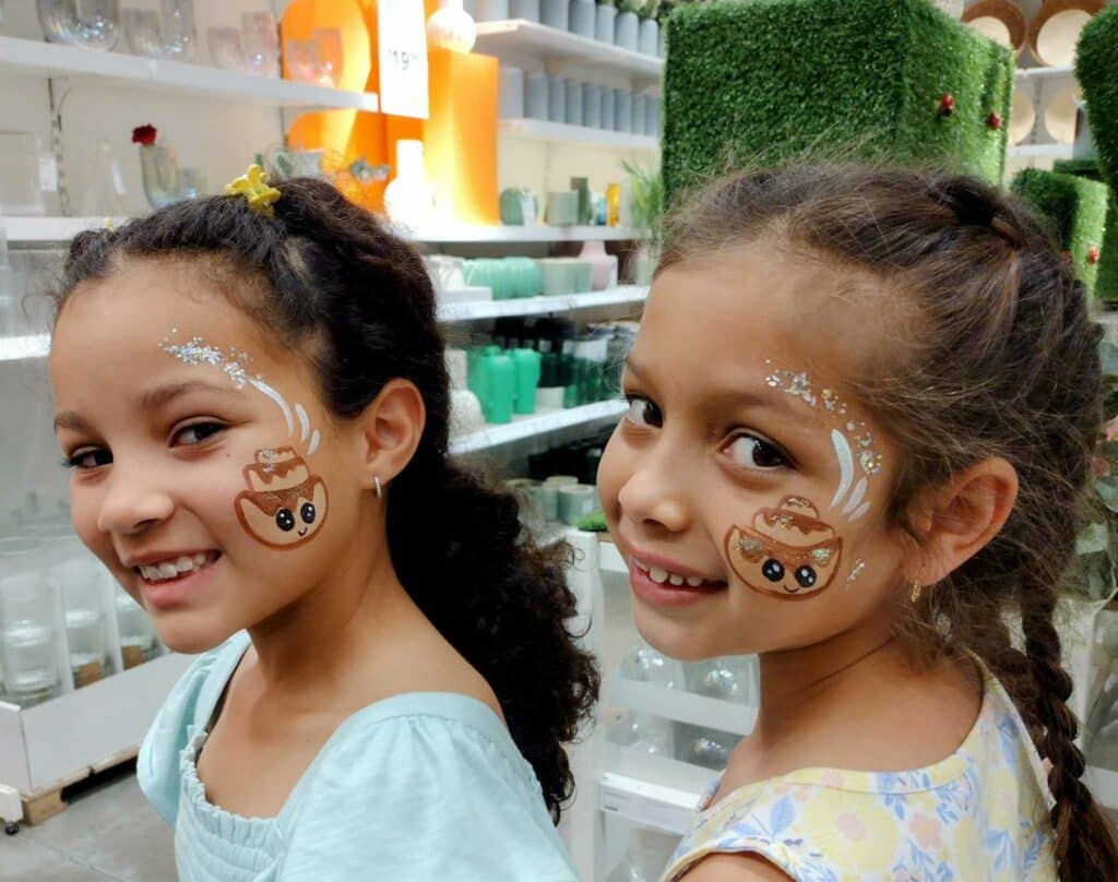Two young girls with face paintings resembling cinnamon rolls on their cheeks, smiling and looking at the camera. They are participating in a fun activity at an IKEA event, with festive decorations in the background. The cheerful scene is captured in a family-friendly environment in Orlando, Florida.