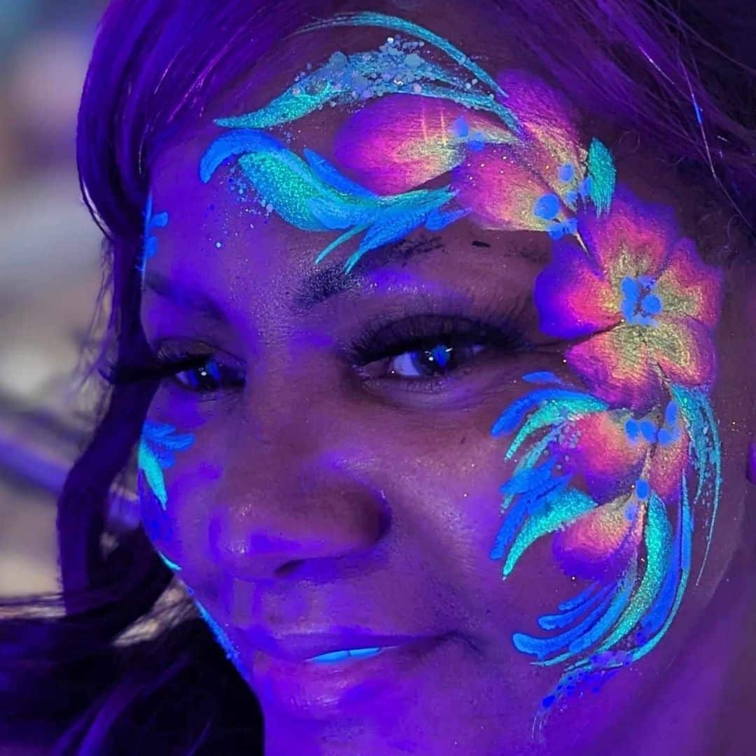 A close-up of a woman with intricate glow-in-the-dark face paint featuring bright, neon floral designs that stand out against the dark background.