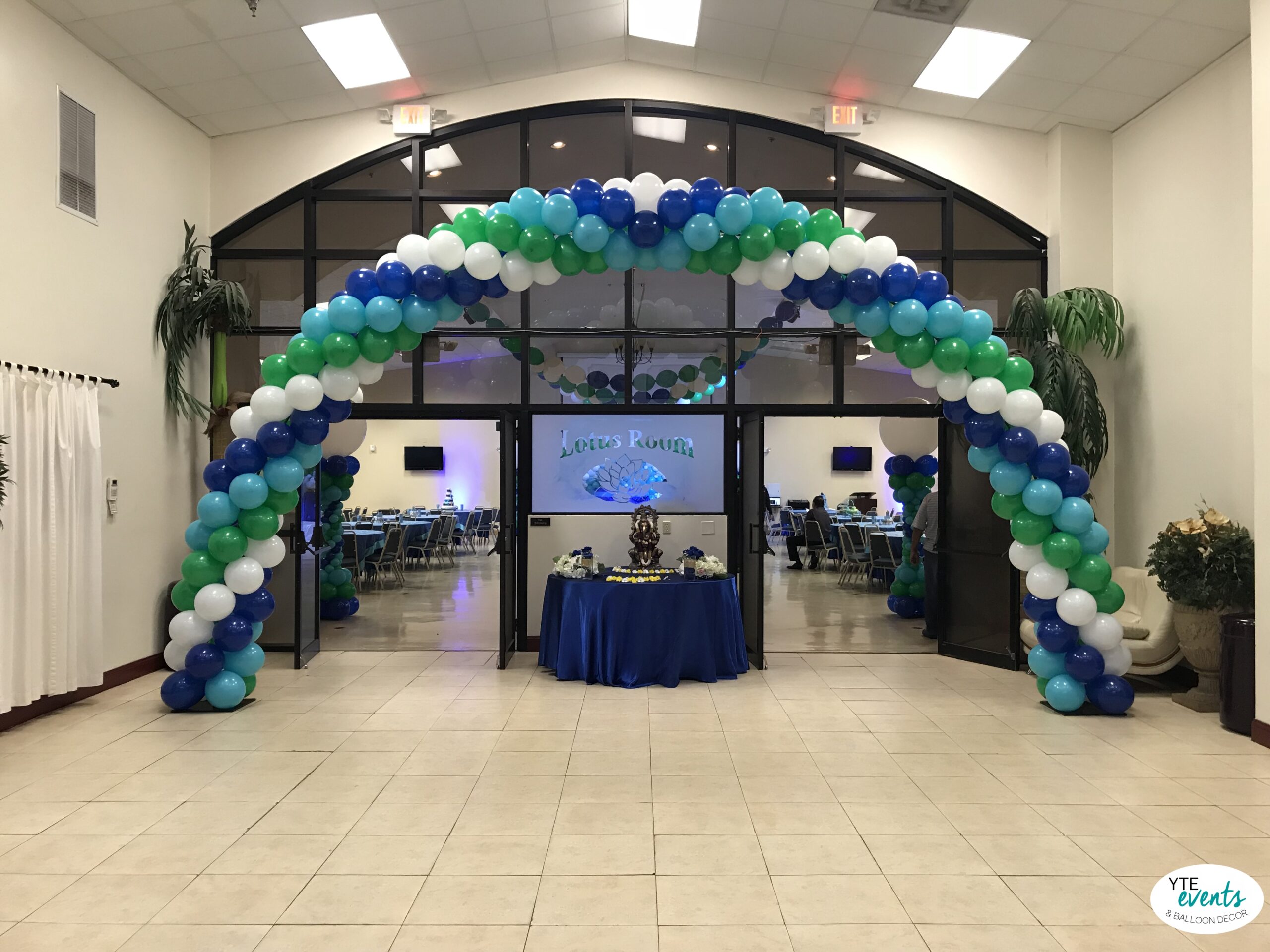 Large balloon arch with green, blue, teal and white balloons at entrance of event room decorated for corporate or social gathering