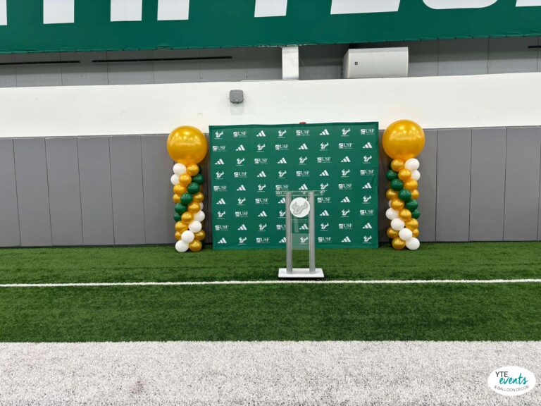 graduation decor Green and gold balloon columns flanking a USF branded stage backdrop with podium