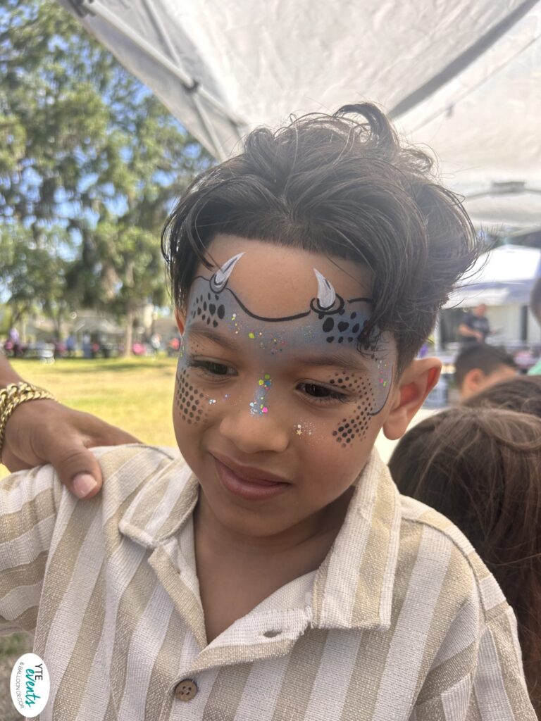Young boy with dragon-themed face paint including horns and glitter outdoors