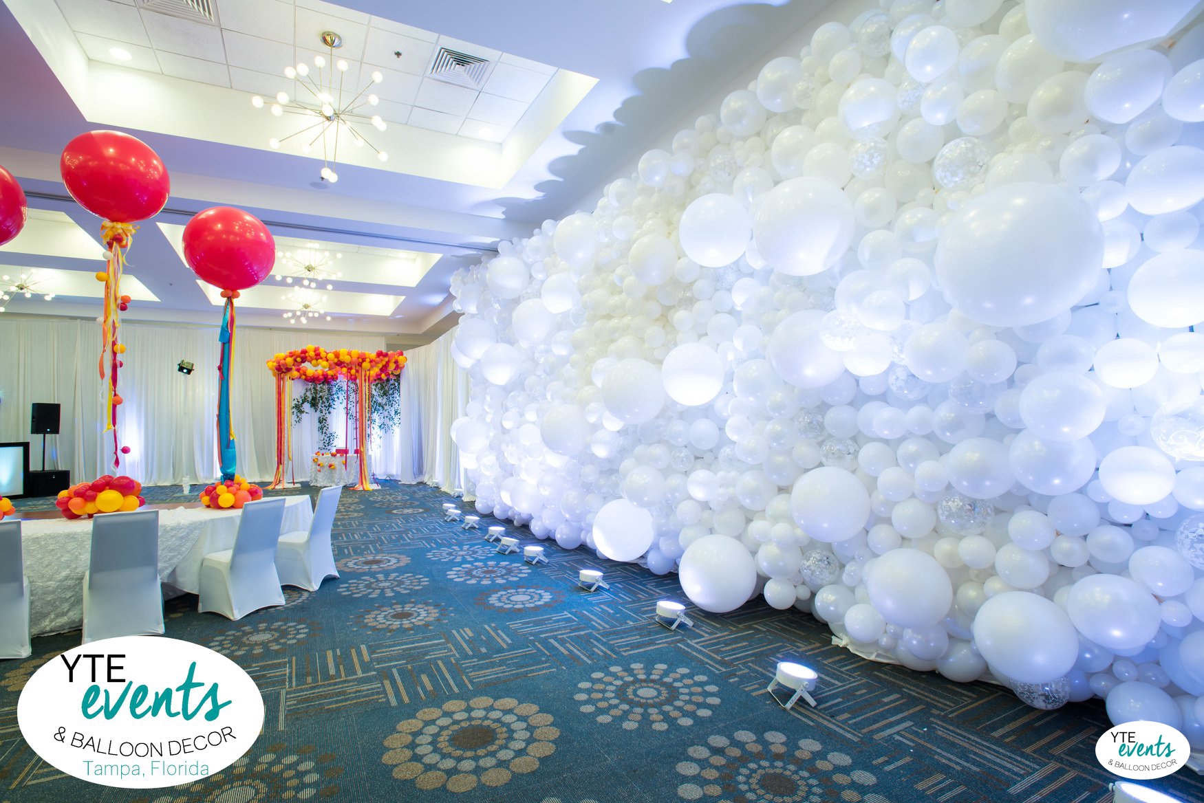Large white balloon wall with varying sizes and pink orange balloon table decorations in a spacious ballroom event setup