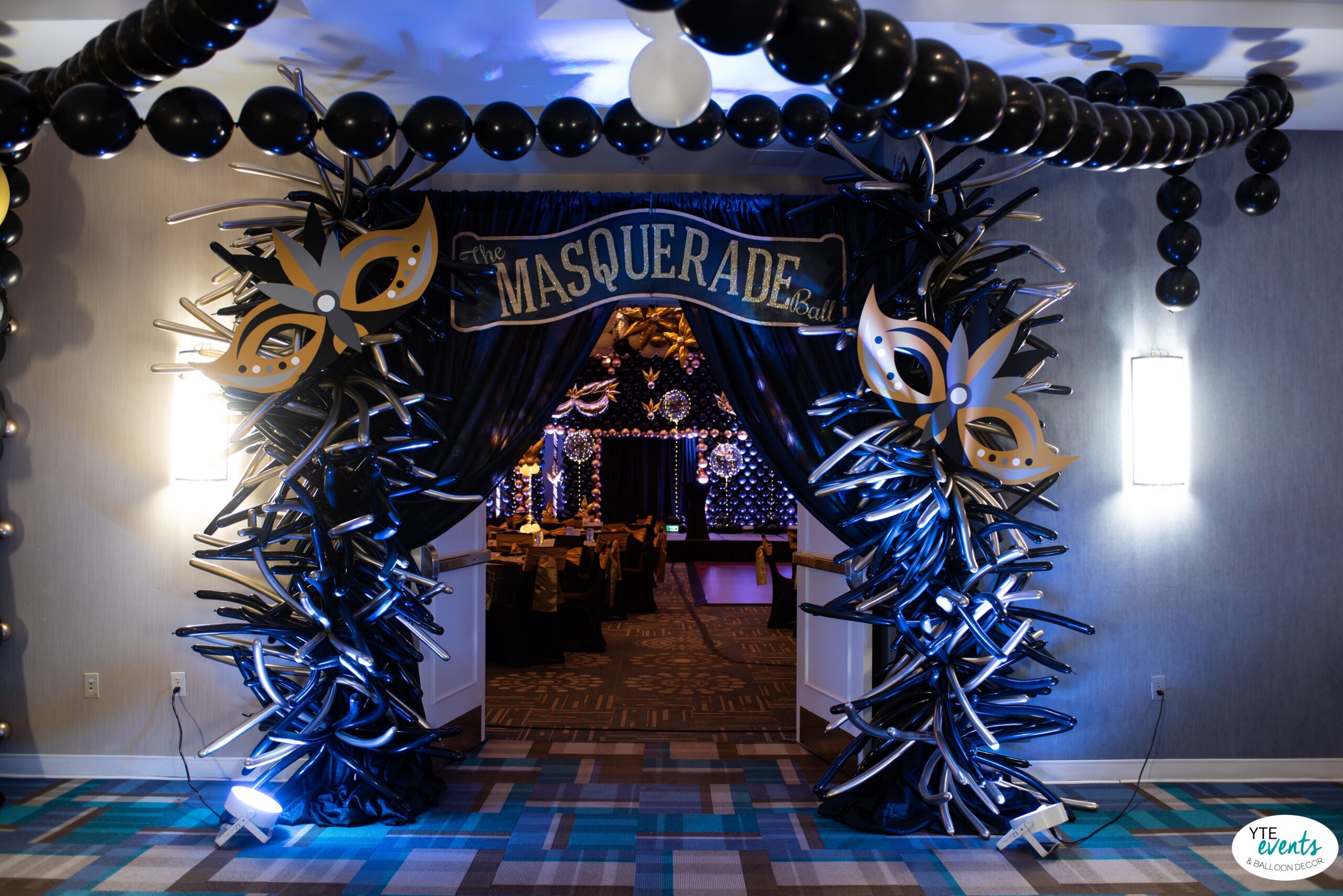 Masquerade ball entrance decorated with black and silver twisted balloon pillars, black balloon garlands, and gold decorative masks