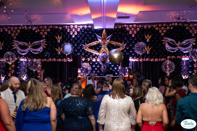 Masquerade ball event with an elaborate black and gold balloon wall featuring balloon masks and clear balloons with LED lights