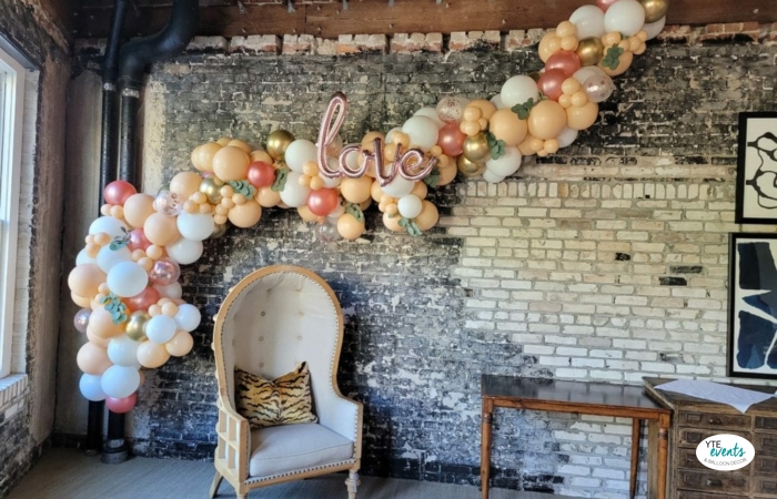 Organic balloon garland with metallic pink, white, peach, and gold balloons forming an arch on a rustic brick wall with a love balloon sign