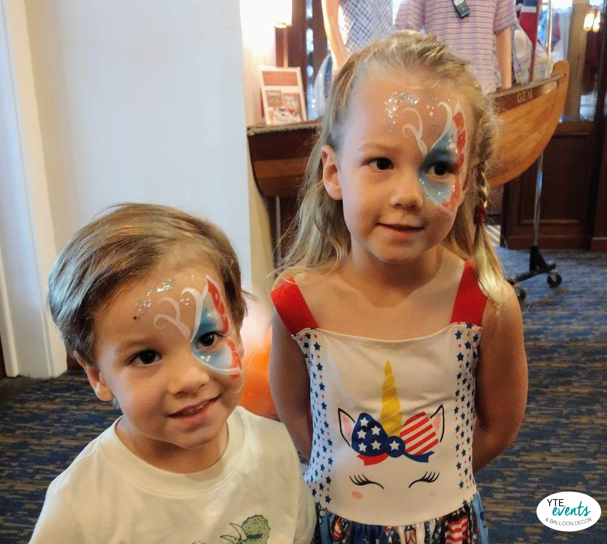 Two young kids with red, white, and blue patriotic face paint designs, smiling indoors