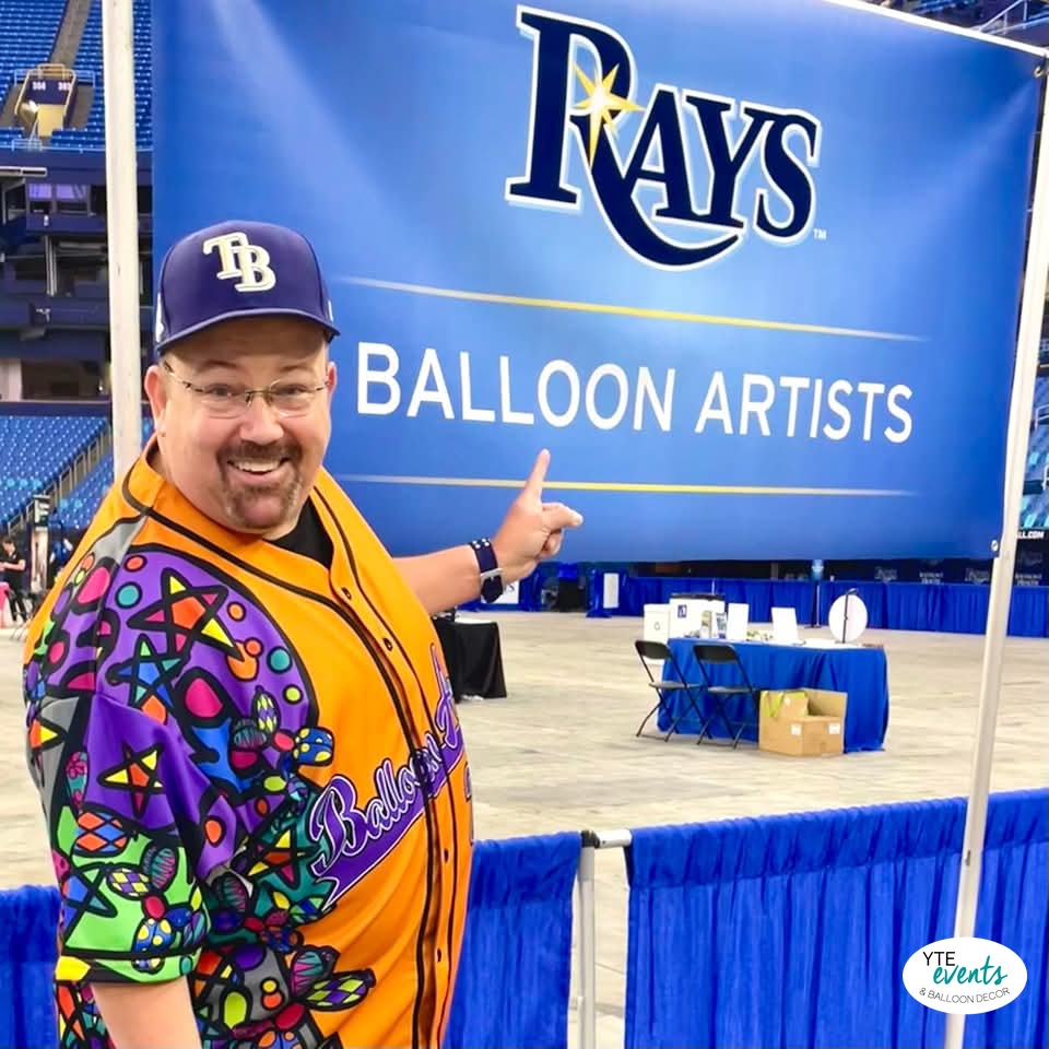 Smiling man in colorful balloon-themed jersey pointing to Rays Balloon Artists banner at indoor corporate event