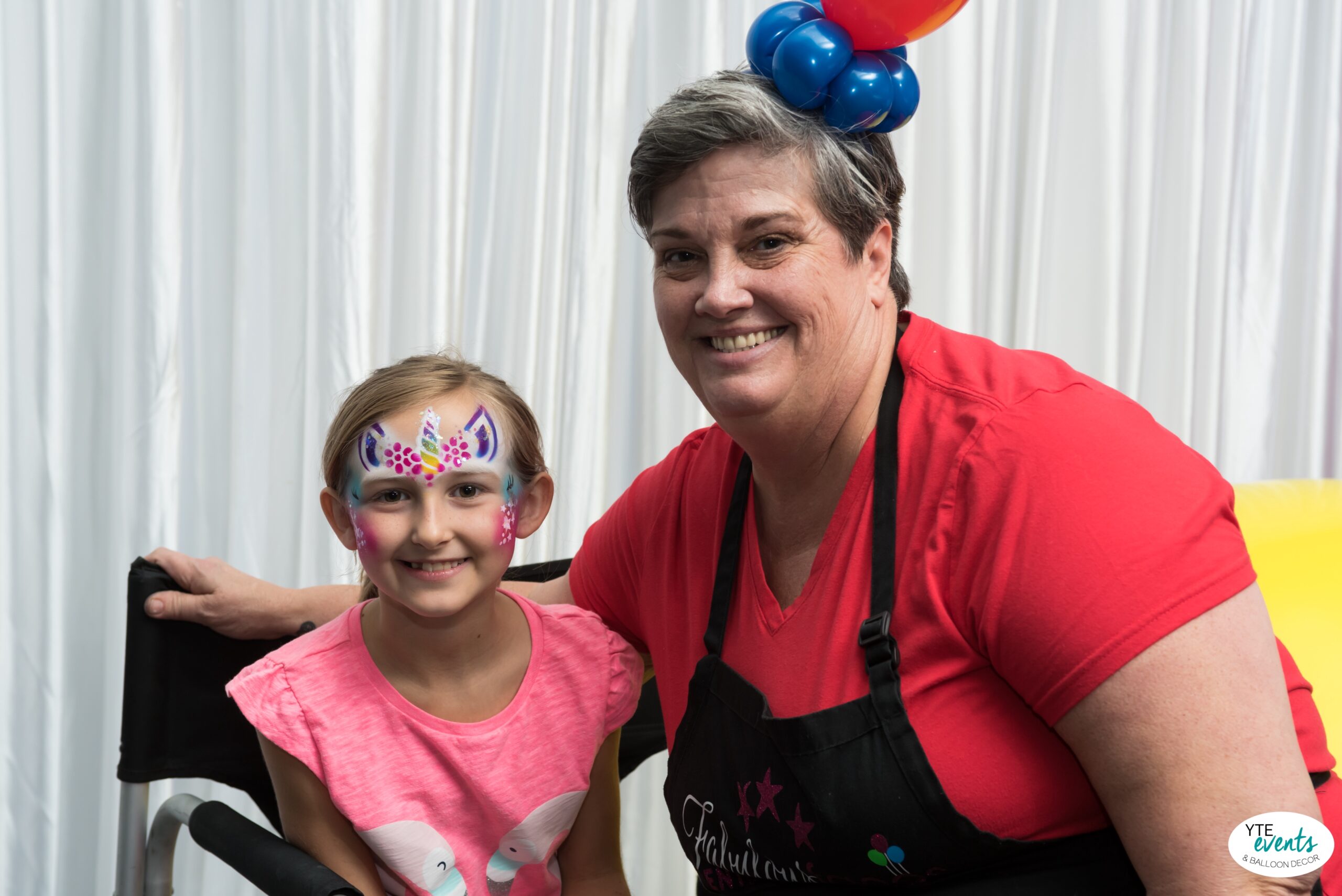 Young girl with colorful unicorn airbrush face painting sitting next to smiling female face painter in red shirt