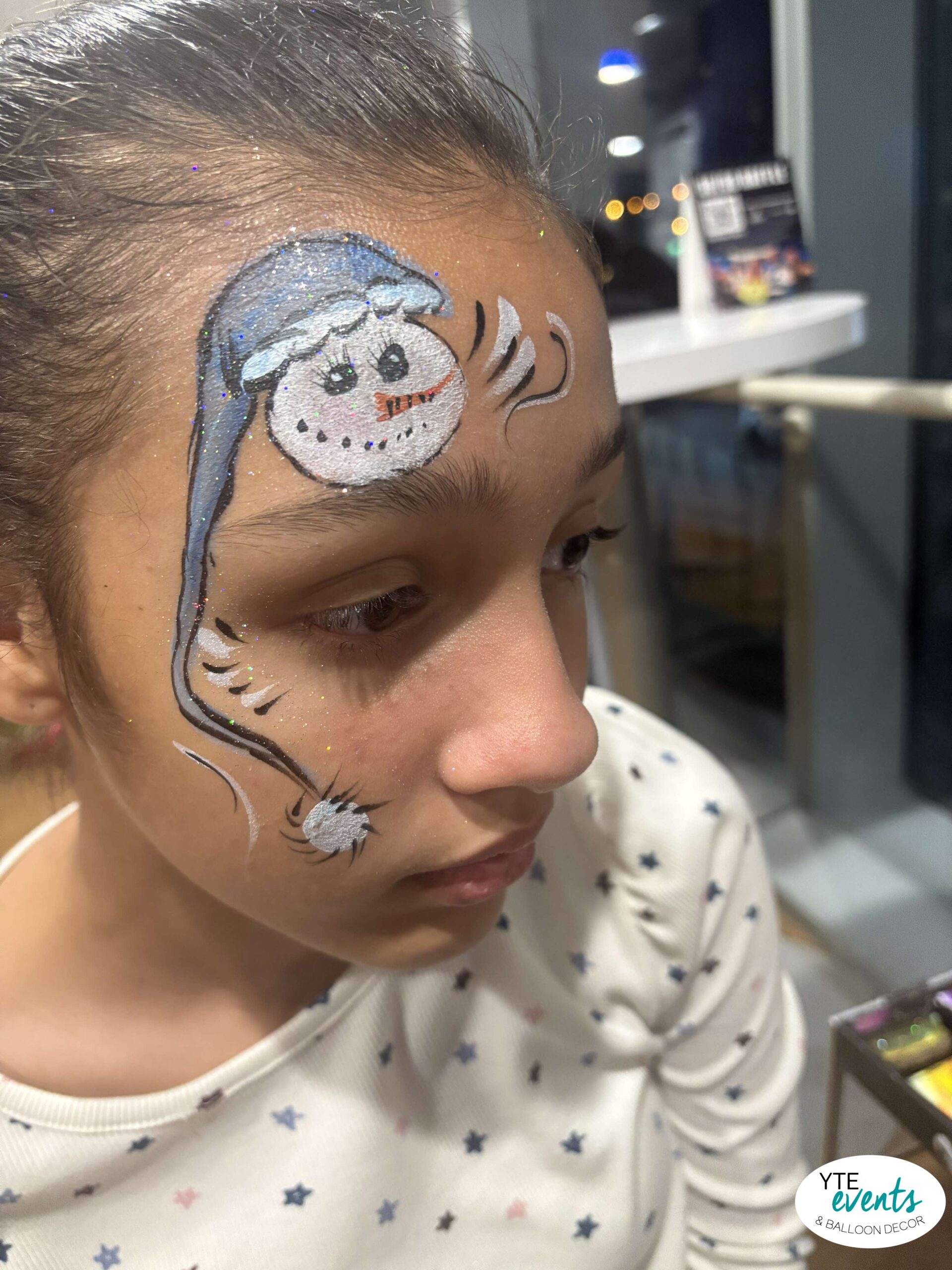 Close-up of a child with glittery snowman face painting featuring blue hat and white snowball details